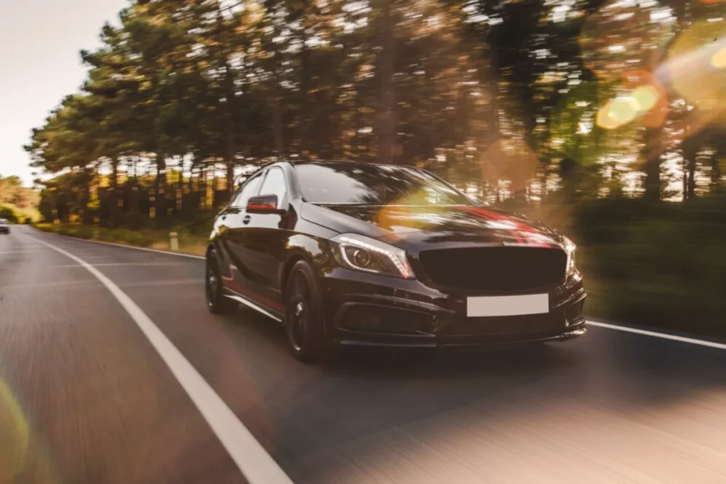 A black sedan speeding on a highway surrounded by trees, with sunlight filtering through the foliage
