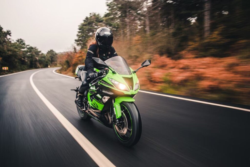 A rider in black gear speeds on a vibrant green motorcycle along a winding road surrounded by autumn foliage.