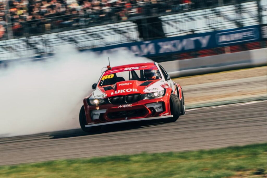 A red BMW drift car with the number 80 skids on a racetrack, surrounded by smoke, while spectators watch in the stands.
