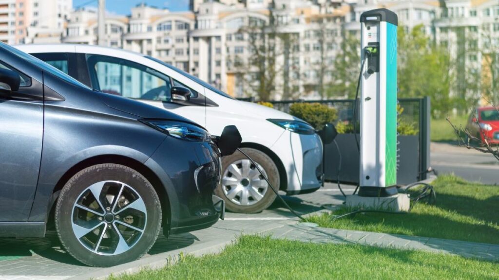 Two electric cars parked at a charging station, one dark gray and one white, with buildings in the background and green grass below.